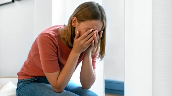 Vrouw - Janou - zit op het bed met haar handen voor haar ogen in een rood shirt met blauwe broek