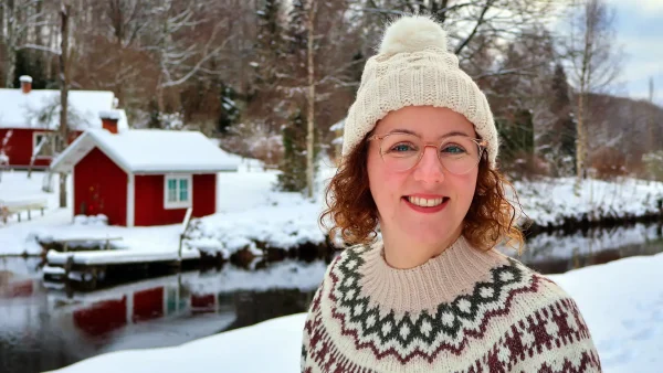 Suzanne staat met een dikke trui en witte muts in de sneeuw in Zweden, met op de achtergrond haar rode huis