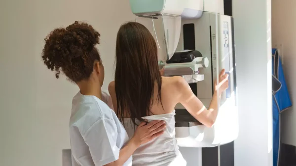 Vrouw neemt plaats voor machine die mammografie zal uitvoeren, onder begeleiding van een arts